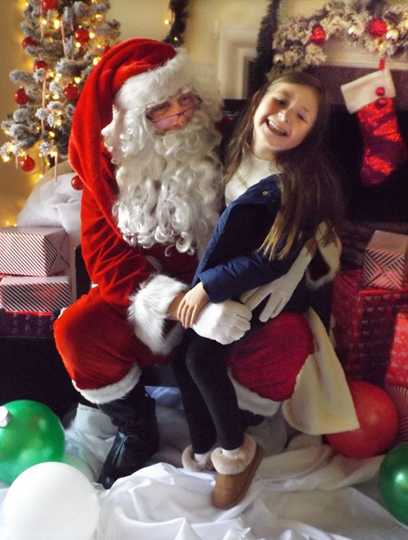 Santa holds a smiling girl beside a decorated Christmas tree, near a  hanging stocking, some gift boxes & other decorations