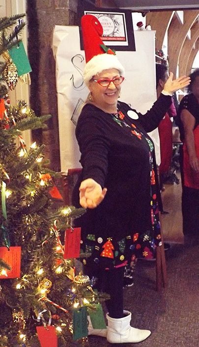 Woman wearing red glasses & a Santa hat poses beside an easel & a decorated Christmas tree, smiling a greeting w/arms raised.