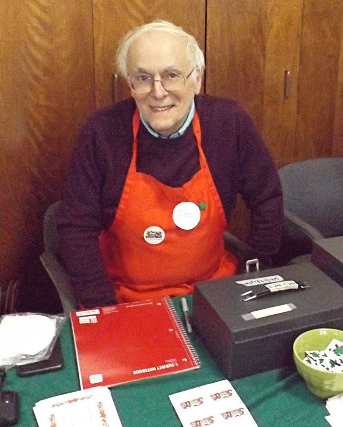 A smiling man in an orange apron sits behind a table, w/a metal cashbox & spiral notebook.
