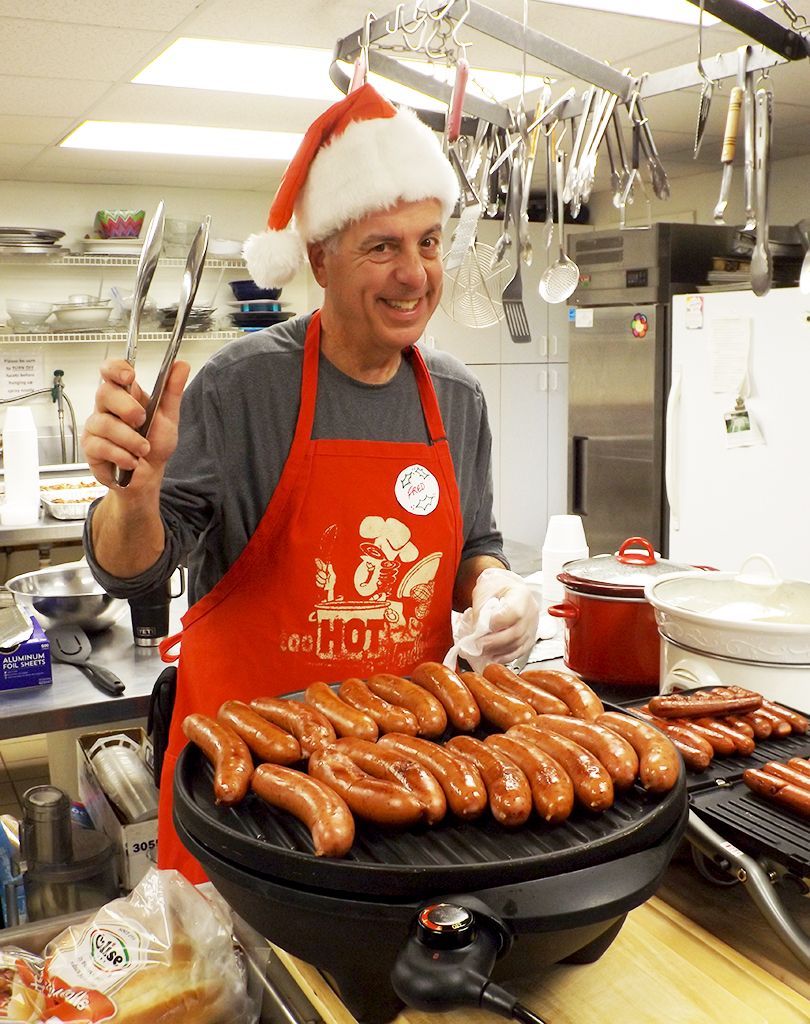 Man in Santa hat & red apron grills kielbasa in a kitchen, smiling beside a stovetop