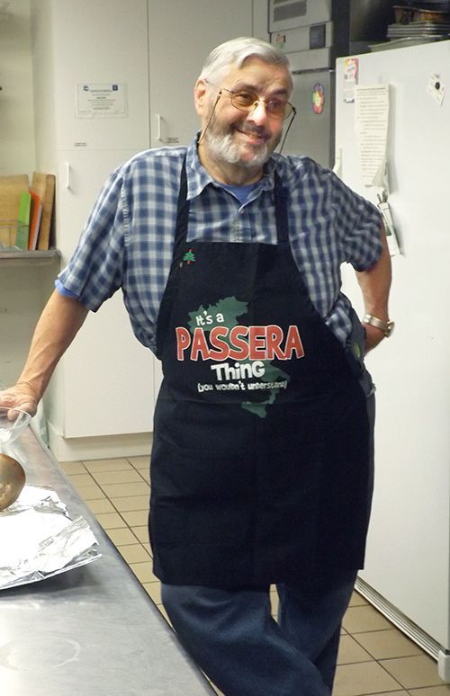 Smiling man in glasses & a black apron stands in a kitchen beside a counter & refrigerator.