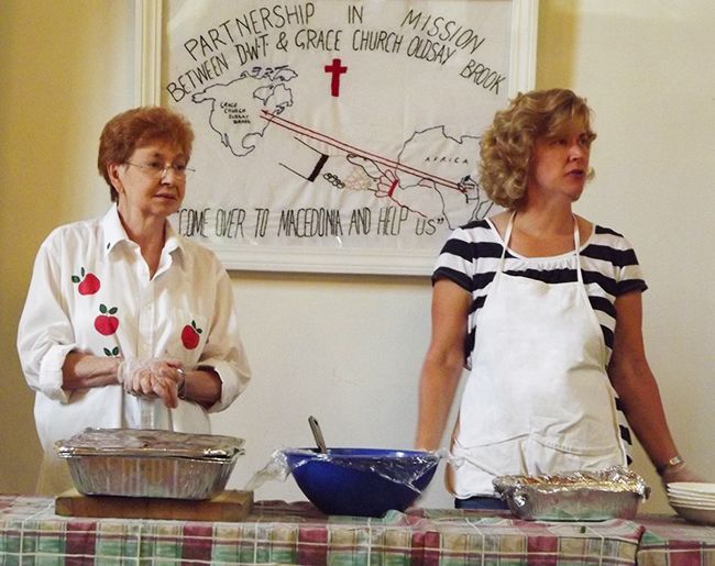 Two women in aprons stand behind serving bowls on a table at an SSKP church luncheon