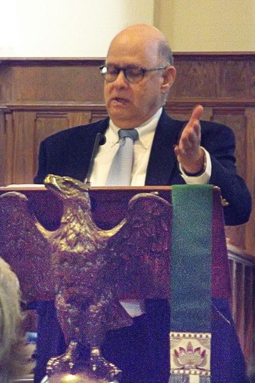 A suited man speaks at church podium with an eagle emblem, as he gestures with one hand.