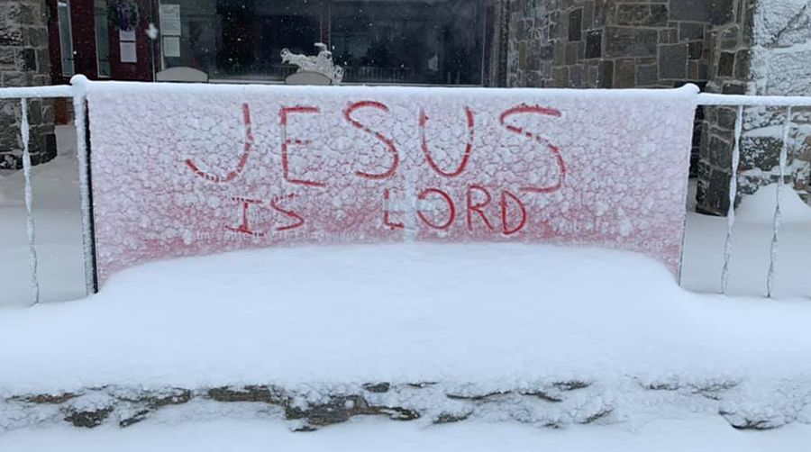 A snow-covered red sign on the church patio has hand-written graffiti in the snow, reading “JESUS IS LORD”