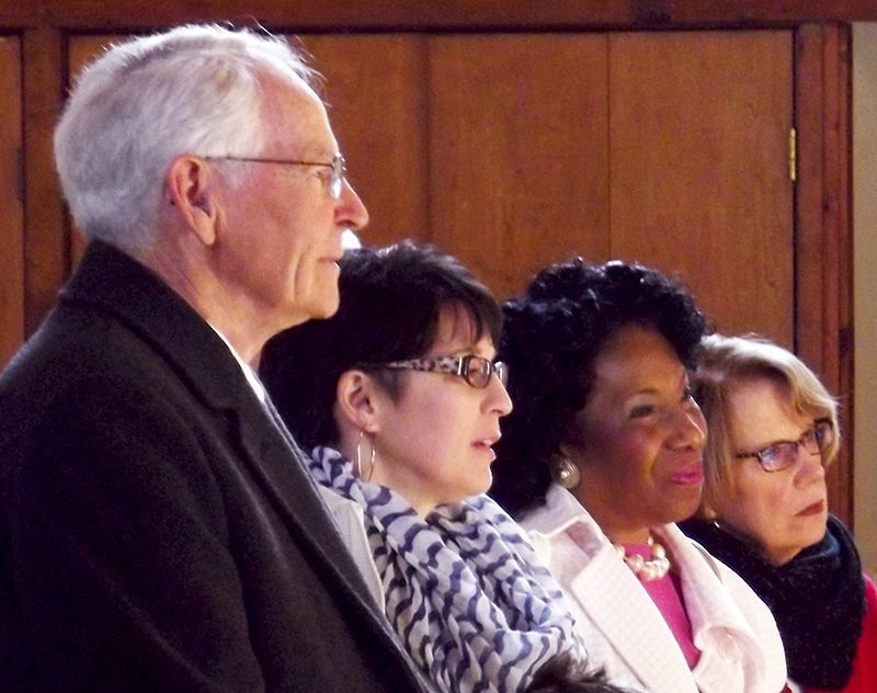 Four people stand in profile at a formal indoor event, with the wood-paneled walls of the Parish Hall behind them.