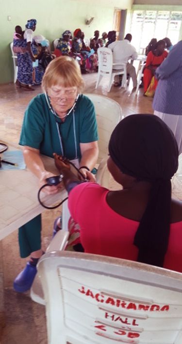 Nurse wearing a stethoscope checks a seated patient’s blood pressure in a busy clinic waiting area.