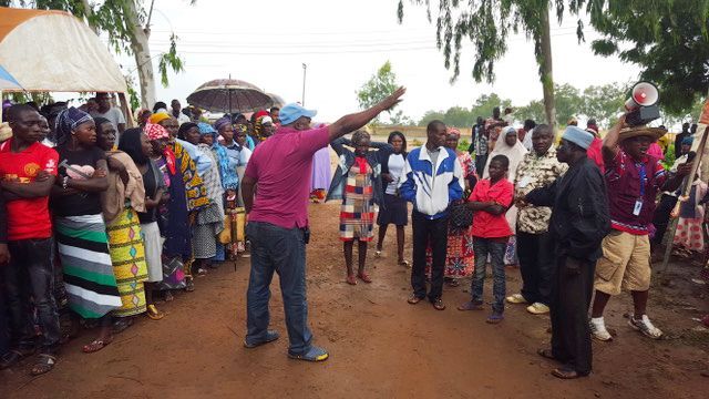 A man raises his hand to address a group of community members gathered outdoors under umbrellas on a muddy path.