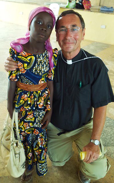 A radiantly beaming Pastor in black clerical attire kneels side by side with a young Black African girl in colorful clothing