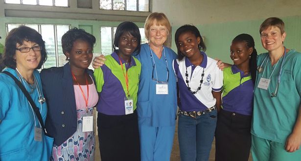 Seven women, some dressed in scrubs, stand smiling together wearing name lanyards indoors, near a window in a clinic