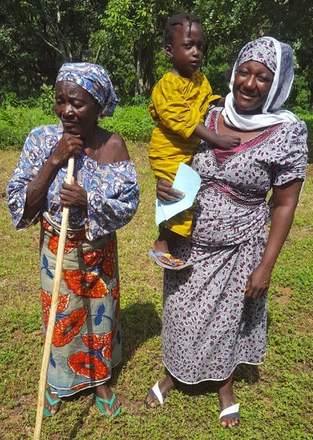 2 women, one holding a child, stand on the grass outdoors, in patterned clothing & headscarves, w/trees & shrubs behind them.