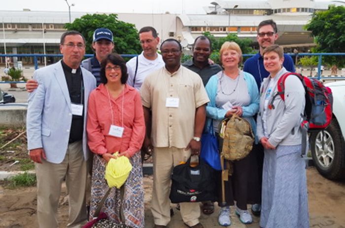 A group of people stand outdoors, in front of a building & parked cars, wearing name tags.