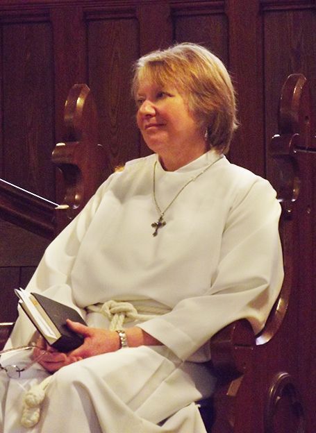 Female Eucharistic Minister in a white robe seated in a wooden church pew, holding a Bible and smiling gently.