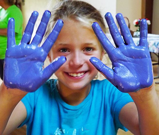 A smiling little girl holds up blue-painted hands from fingerpainting.