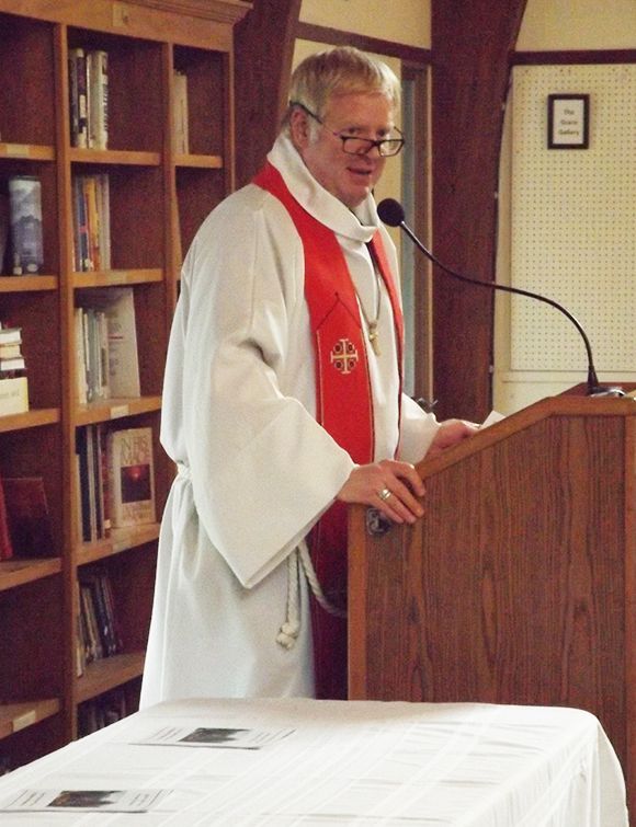 Pastor Charlie in white robes & a red stole speaks at the microphone on a wooden lectern in the Parish Halll.