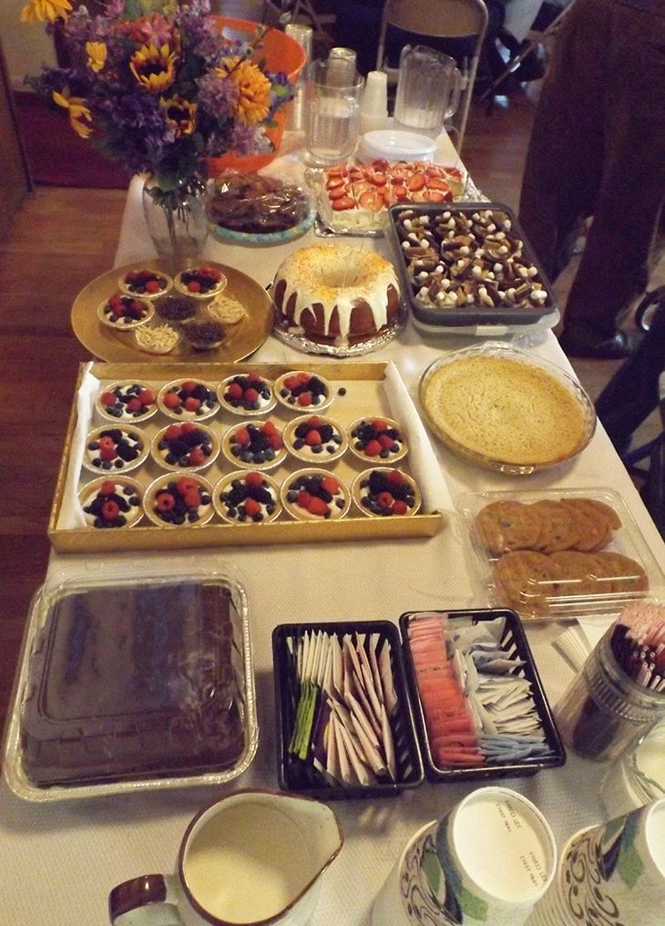 Dessert buffet has assorted cakes, tartlets, pastries, utensils & flowers displayed on a table.