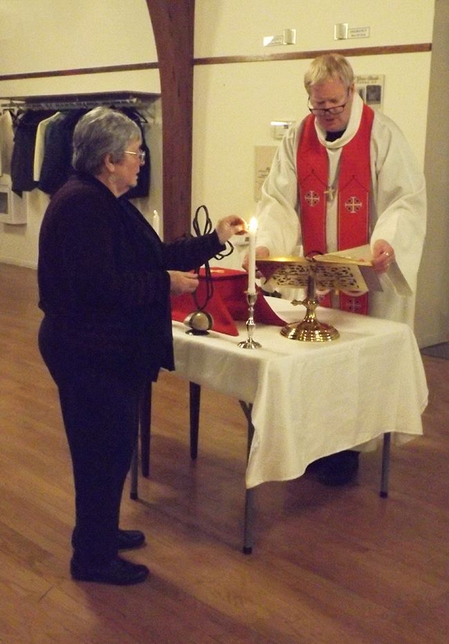 A woman lights candles at a small makeshift altar, while Pastor Charlie stands before her in white & red vestments.