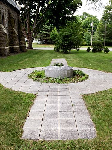 A stone sundial is set into a circular stone pathway on the church's grassy lawn, with trees & shrubs all around.