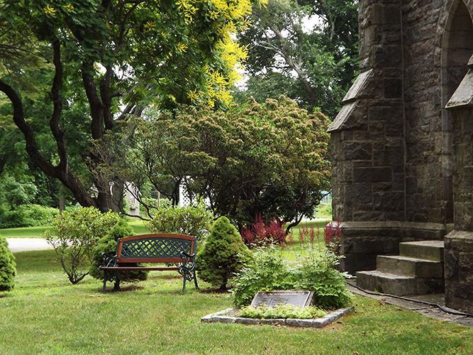 A park bench sits in a lush Memorial Garden w/blooming pink astilbe plants, beside the stone church building & steps.