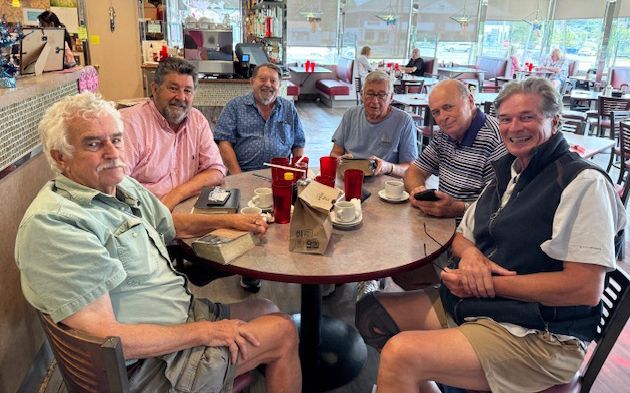 6 men seated around a table, smiling & chatting in a bright restaurant by the windows