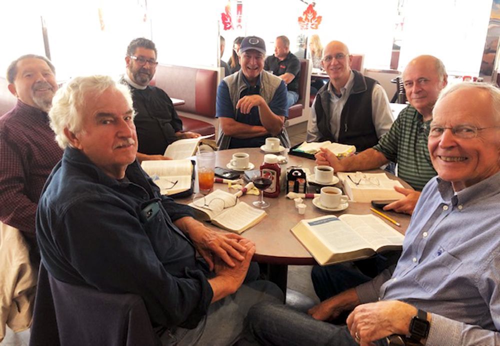 A group of men sits around a table with their Bibles open, smiling & chatting over coffee & beverages.