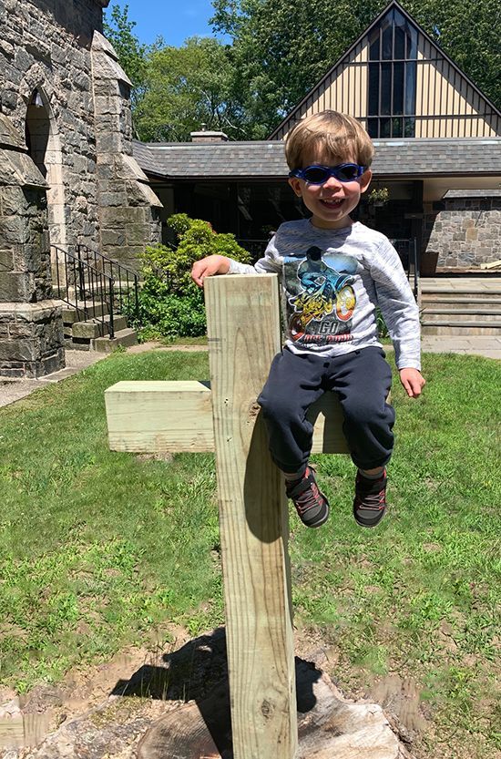 A young boy in sunglasses sits on a wooden cross outdoors near the stone church building