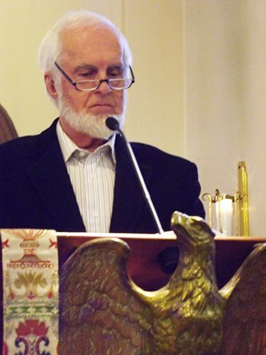 Older man in a dark suit wearing spectacles speaks at a church lectern with a microphone.