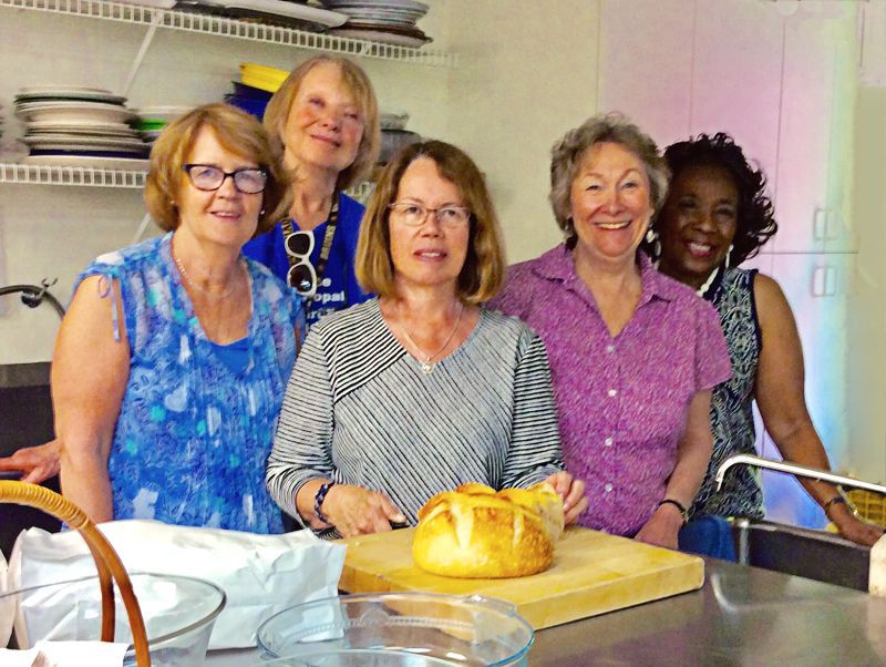 Five women smile behind a kitchen counter with a loaf of bread on a cutting board
