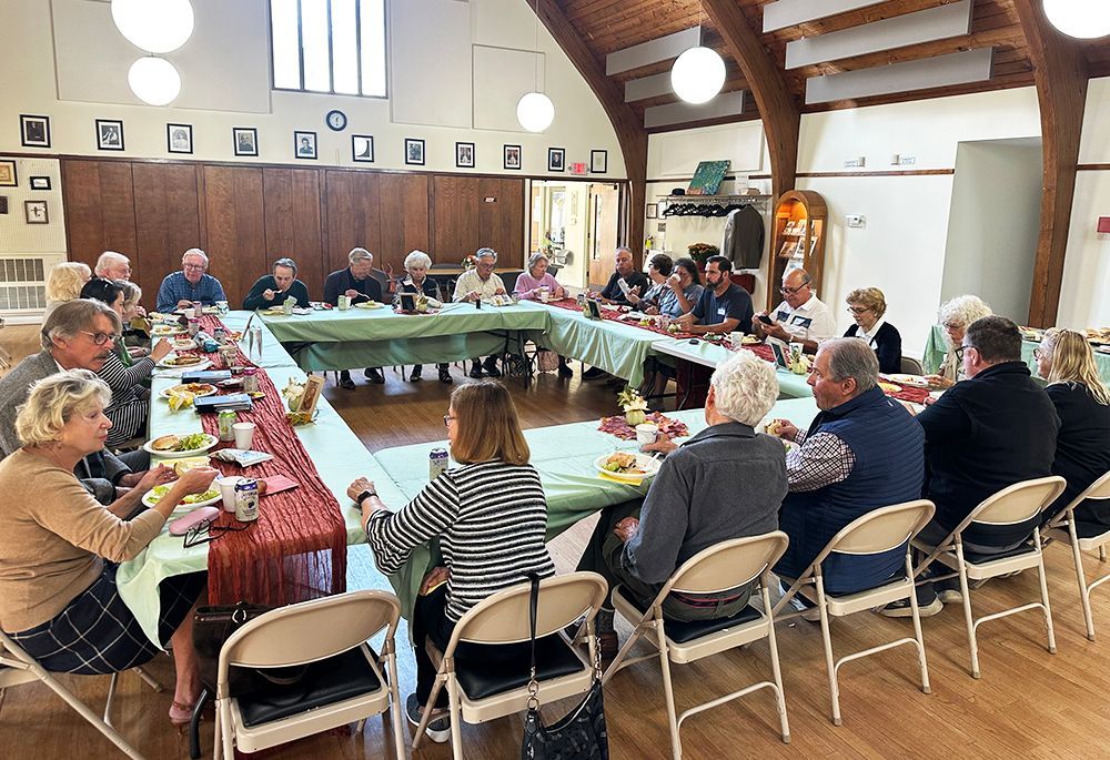 People seated around a long U-shaped table in the bright Parish Hall, eating & discussing together
