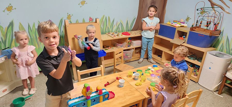 Children play with toys & various colors of clay, in a bright classroom, surrounded by busy shelves & tables.