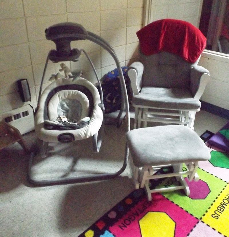 A baby swing & rocker draped in a red blanket sit in a dimmed nursery room w/a gray ottoman nearby.