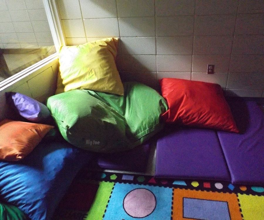 Colorful beanbag cushions lie stacked in a corner, w/purple floor mats and a children’s play rug.