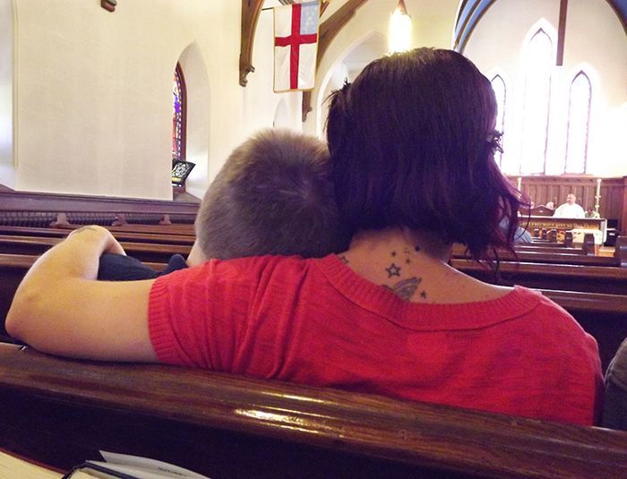 A mother with a star tattoo on her upper back gently curls an arm around her son while sitting in church pew facing the altar