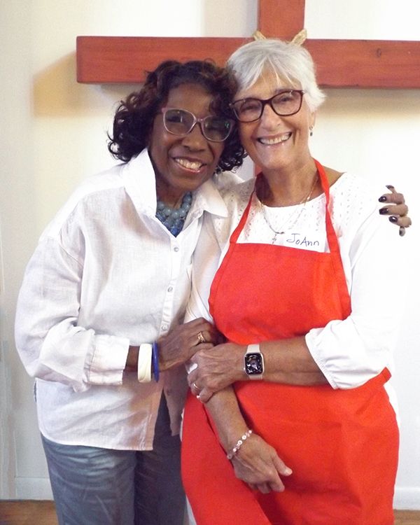 Two smiling women wearing glasses stand close together in front of a red wooden cross, one wearing a red apron.