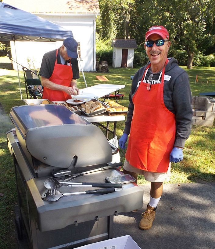 Two men in orange aprons prepare food at a grill during an outdoor picnic with a table and tent nearby.