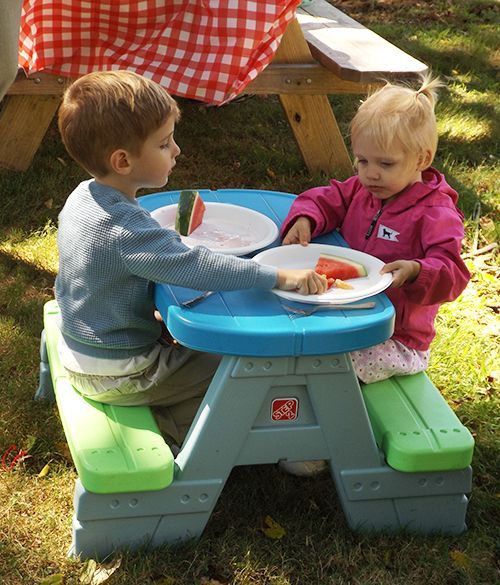 Two toddlers sit at a blue & green picnic table, eating watermelon slices outdoors in the grassy churchyard.