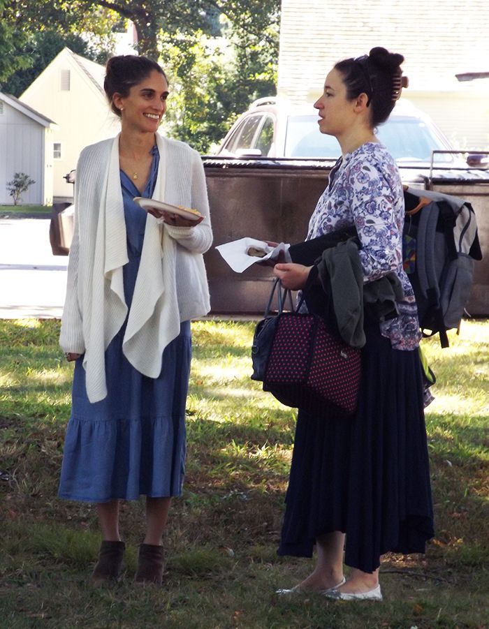2 women chat outdoors in the grassy back churchyard, holding papers & wearing long skirts