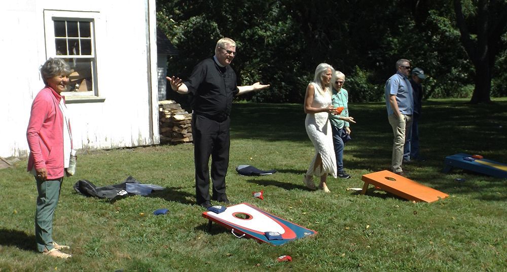Pastor Charlie &some parishioners play cornhole-games outdoors on the grassy churchyard lawn, beside a white shed.