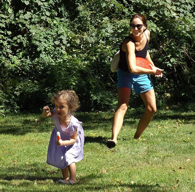 Mother prepares to throw a frisbee as her toddler runs across the grassy lawn with dense green bushes in the background.