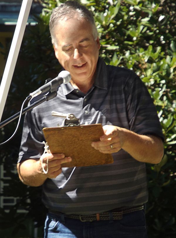 A man reads from a clipboard into a microphone outdoors in the sunlight