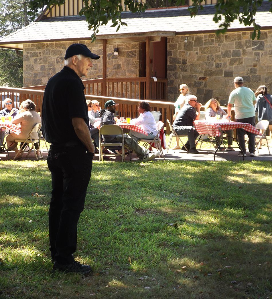 Man in black stands on grass in front of a stone building, with people seated at picnic tables behind him