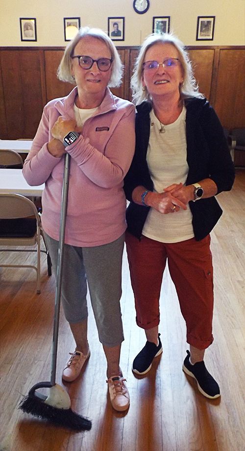 Two women stand in the Parish Hall for cleanup, one holding a mop, with framed pictures on the wall behind them.