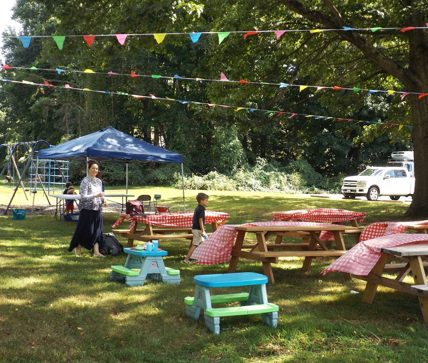 Outdoor picnic area has red-checked tables, a blue canopy, colorful hanging pennants, & a white truck in the background