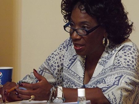 A female intake-worker wearing glasses, jewelry & a patterned blouse, speaks at a table.