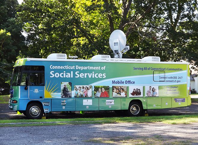 CT Department of Social Services Mobile Office bus, parks outdoors in the church parking lot under leafy green trees.