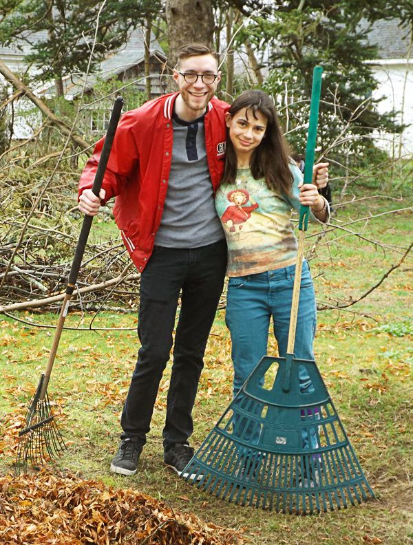 A young man & woman stand side-by-side outdoors holding garden tools near some piles of brush & leaves.