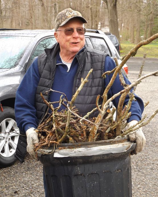 Man wearing a cap, fleece jacket, vest & work-gloves carries a trash bin holding a bundle of branches next to a parked SUV 
