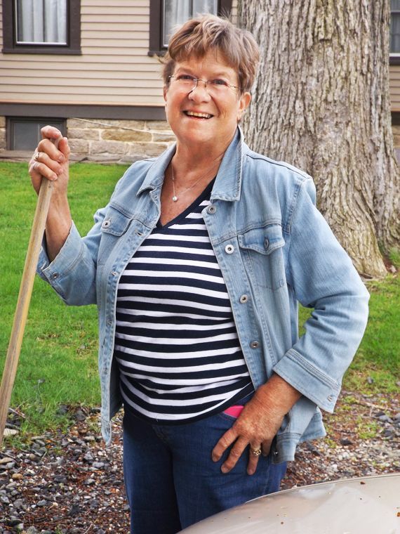 A smiling woman wearing a denim jacket & striped shirt stands outdoors holding a rake next to a tree.
