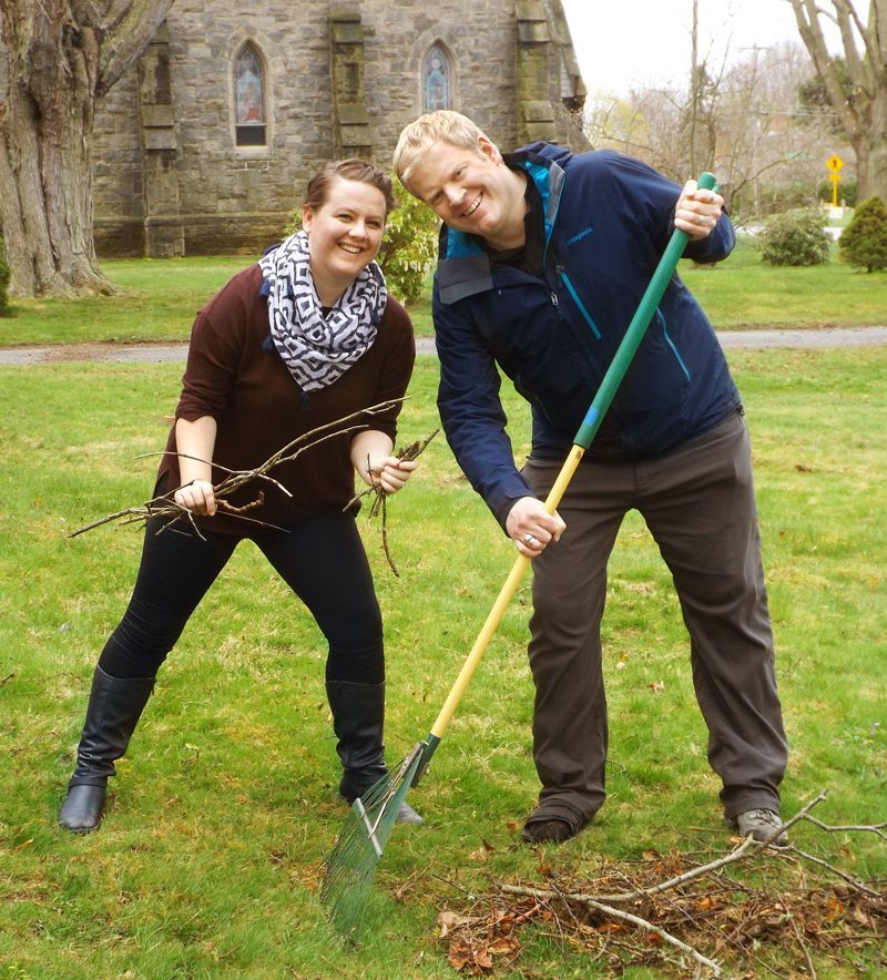 Pastor Charlie stands w/a female parishioner, raking leaves on a grassy lawn in front of the stone church