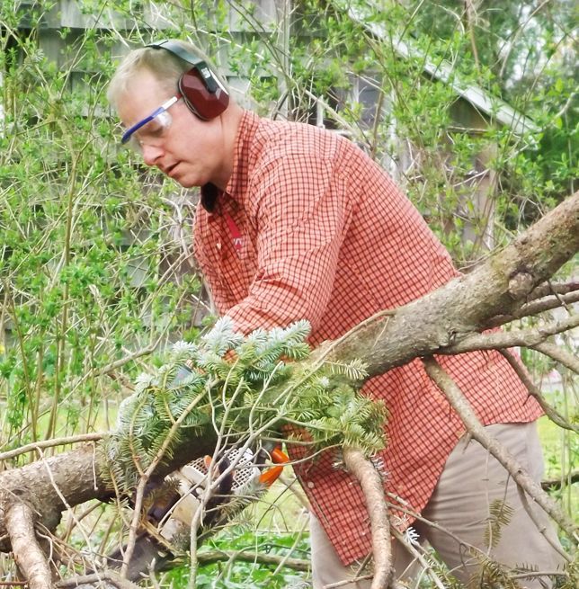 A man wearing an orange shirt, safety glasses & ear protectors cuts a fallen tree branch outdoors with a chainsaw.