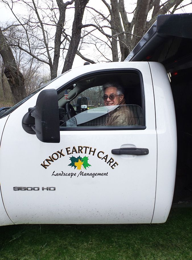 A male driver wearing sunglasses in a white landscaping truck, smiles while parked on the grass near bare trees